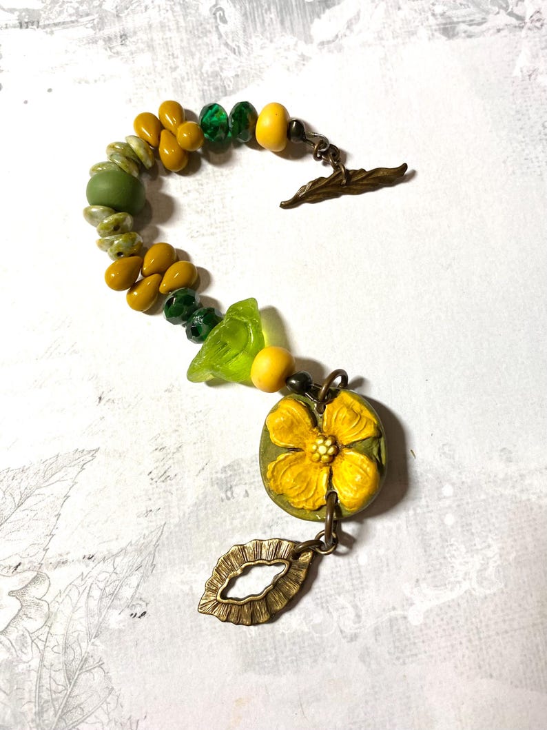 Puede incluir: Un brazalete artesanal con una mezcla de cuentas amarillas, verdes y transparentes. El brazalete incluye un dije de flor amarilla detallada y un cierre en forma de hoja de bronce. Las cuentas son de varias formas y tama&ntilde;os.