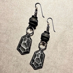 May include: A pair of black and silver earrings with geometric shapes and textured details. The earrings feature a black bead with silver accents and a black pendant with a textured surface.