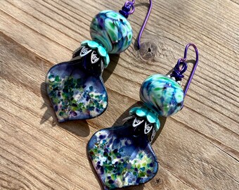May include: A pair of dangle earrings with a blue and green swirl glass bead and a silver flower charm. The earrings are made with purple wire.