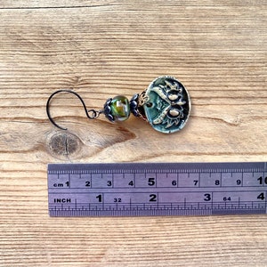 May include: A green and gold ceramic earring with a floral design and a black wire hook. The earring is about 2 inches long.