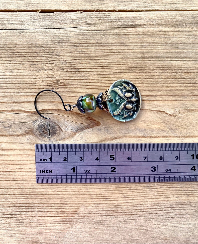May include: A green and gold ceramic earring with a floral design and a black wire hook. The earring is about 2 inches long.