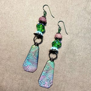 May include: A pair of dangle earrings with a green, pink, and blue color scheme. The earrings feature a teardrop-shaped pendant with a wave pattern and are accented with glass beads.