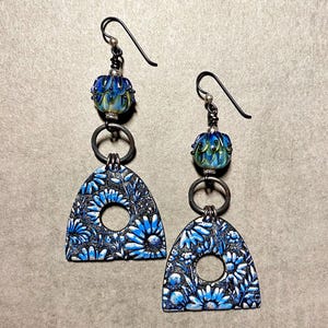 May include: A pair of dangle earrings with a blue and green glass bead and a silver charm with a floral design. The charm is shaped like an arch with a circle in the center.