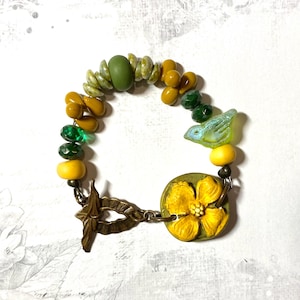 Puede incluir: Una pulsera hecha a mano con cuentas verdes, amarillas y doradas. Incluye un peque&ntilde;o dije de p&aacute;jaro y un dije redondo con un dise&ntilde;o de flor amarilla. El cierre es de metal color bronce. Aproximadamente 18 cm de largo.