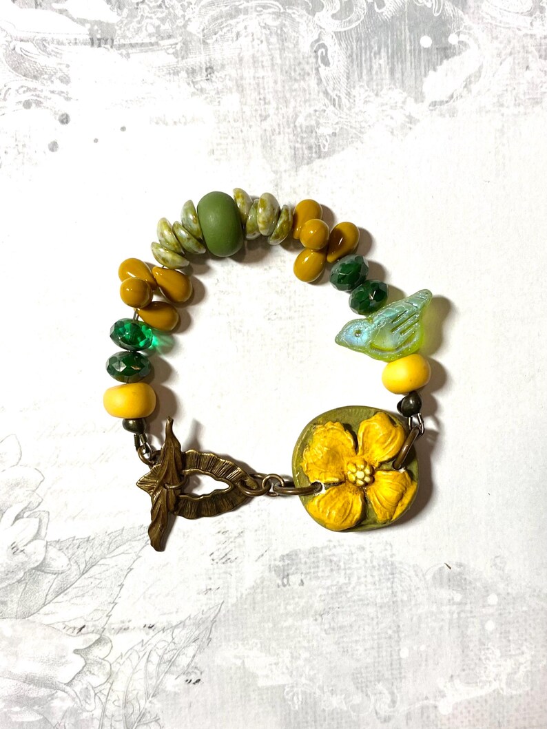 Puede incluir: Una pulsera hecha a mano con cuentas verdes, amarillas y doradas. Incluye un peque&ntilde;o dije de p&aacute;jaro y un dije redondo con un dise&ntilde;o de flor amarilla. El cierre es de metal color bronce. Aproximadamente 18 cm de largo.