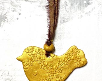 May include: A gold-colored bird-shaped ornament with embossed floral and heart details. It hangs from a brown ribbon and a wooden bead. The bird has a textured surface and is likely a decorative item.