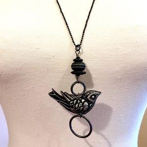 May include: A black necklace with a silver bird pendant and a black and white bead. The bird is detailed with silver accents and has a small eye.