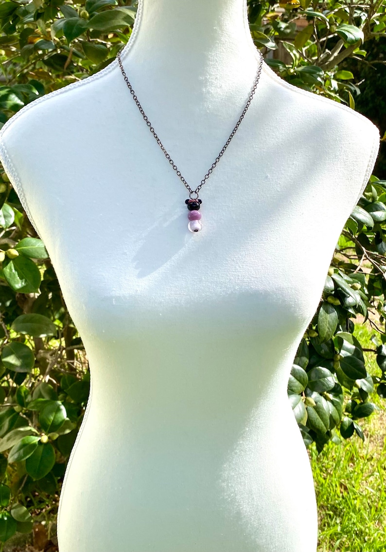 May include: A silver chain necklace with a pendant featuring three beads. The top bead is black, the middle bead is purple, and the bottom bead is white.