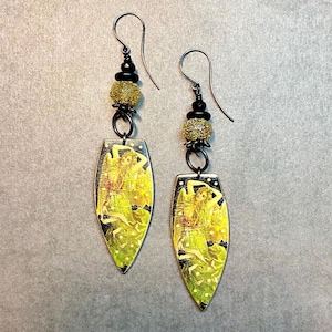 May include: A pair of dangle earrings with a gold-toned, leaf-shaped pendant featuring a vintage illustration of a woman in a green dress. The pendant is accented with black and gold beads.