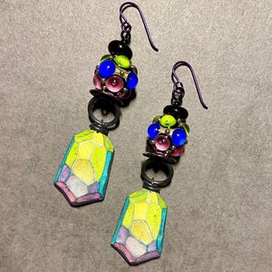 May include: A pair of colorful earrings with a geometric design. The earrings feature a cluster of small, colorful glass beads and a larger, geometric pendant. The pendant is made of a yellow, green, and purple material and has a textured surface.