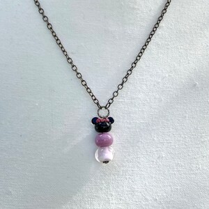 May include: A black and purple beaded necklace with a black mouse charm with a pink bow. The necklace is on a white fabric background.