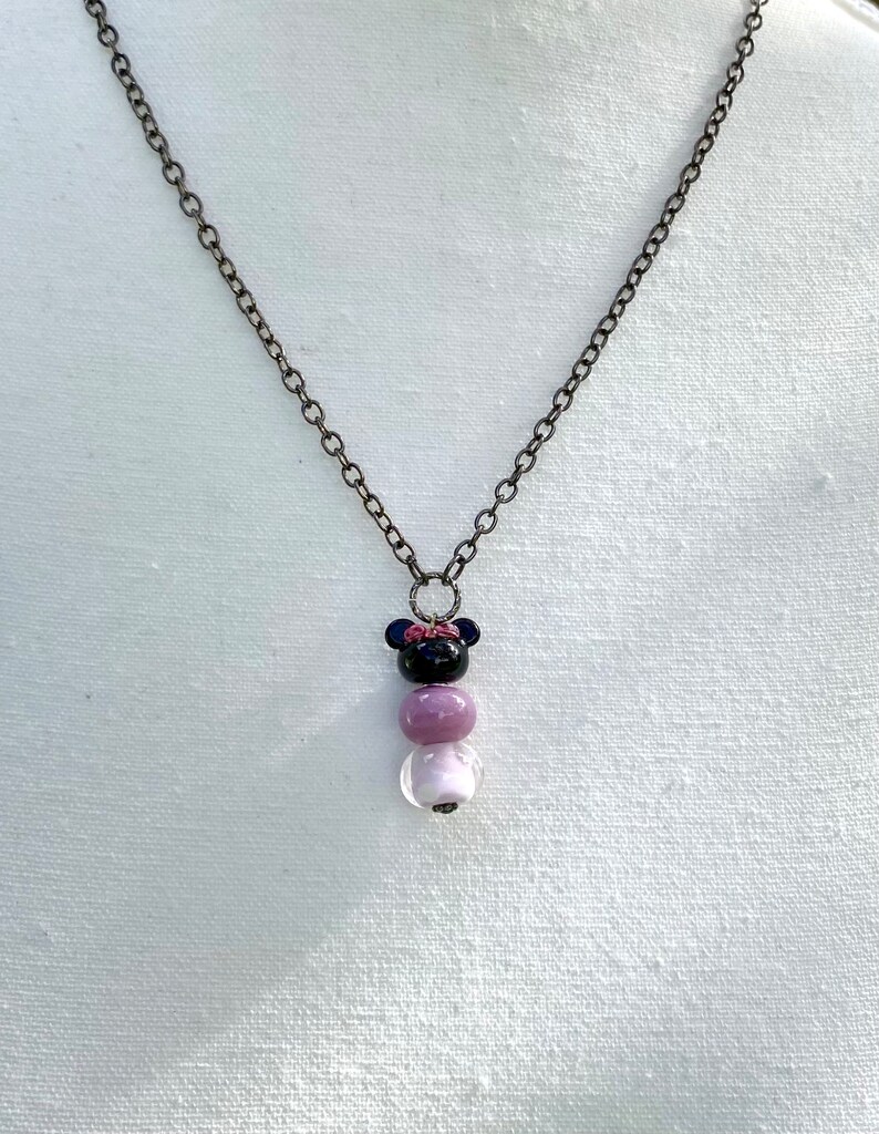 May include: A black and purple beaded necklace with a black mouse charm with a pink bow. The necklace is on a white fabric background.