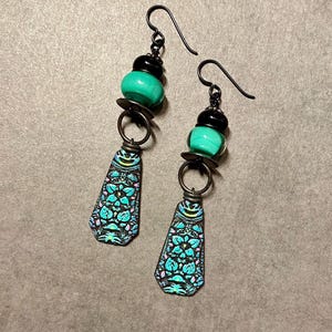 May include: A pair of dangle earrings with turquoise and black beads and a patterned metal charm. The charms are shaped like a teardrop and have a floral design.