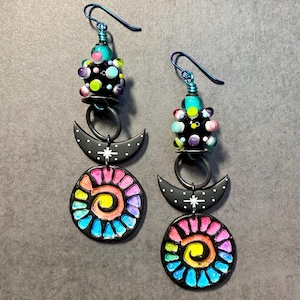 May include: A pair of dangle earrings with a black crescent moon, a colorful sunburst design, and a black bead with colorful dots.