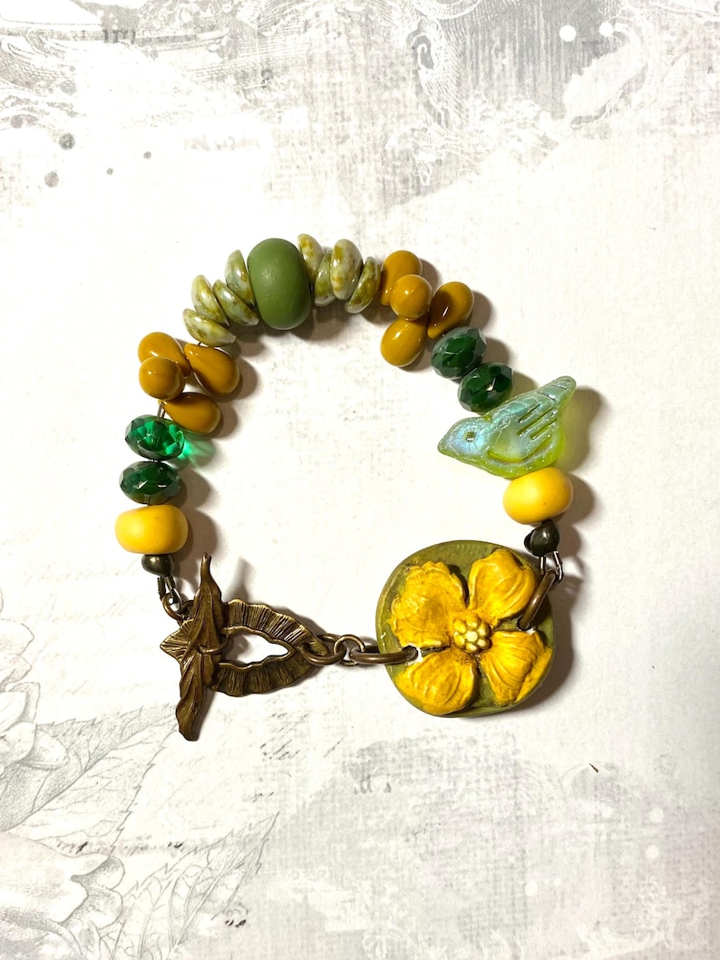 Puede incluir: Una pulsera artesanal con una variedad de cuentas en tonos verdes, amarillos y dorados. La pulsera incluye una cuenta en forma de p&aacute;jaro y un cierre en forma de flor. La pulsera mide aproximadamente 17.78 cm de largo.