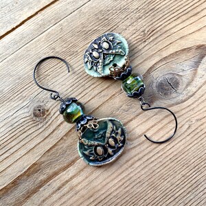 May include: A pair of green and gold earrings with a floral design and green glass beads. The earrings are on a wooden surface.