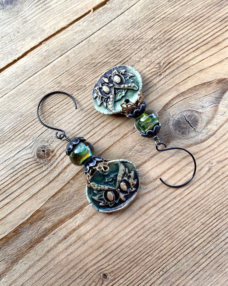 May include: A pair of green and gold earrings with a floral design and green glass beads. The earrings are on a wooden surface.