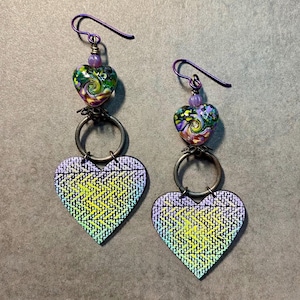 May include: A pair of dangle earrings with a purple, green, and yellow heart-shaped charm. The charm is made of a woven fabric with a metallic sheen. The earrings are attached to a purple wire with a small heart-shaped bead.