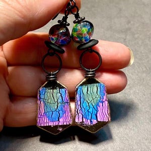May include: A pair of dangle earrings with iridescent glass beads and black metal wire. The beads are hexagonal and have a cracked, textured surface. The earrings are a mix of blue, green, and purple.