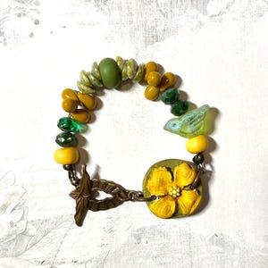 Puede incluir: Pulsera artesanal con cuentas amarillas, verdes y oliva. Incluye un dije de flor amarilla y una cuenta con forma de p&aacute;jaro. El cierre es de metal color bronce.