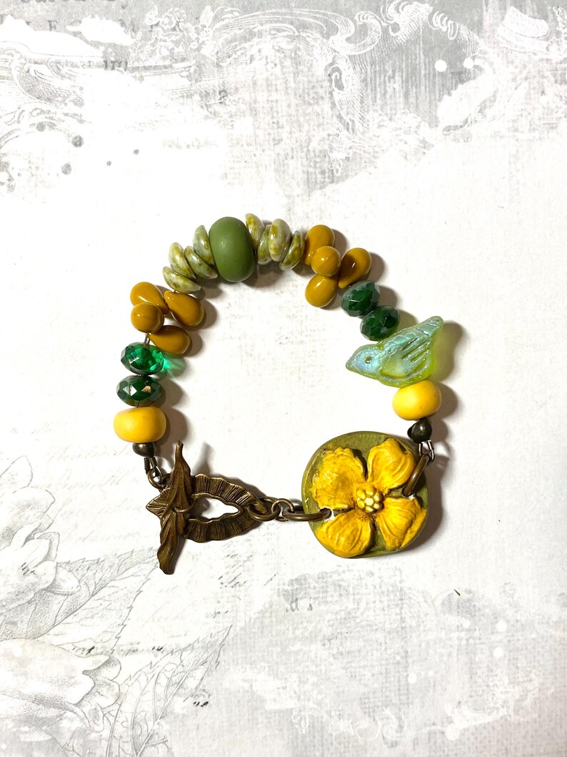 Puede incluir: Pulsera artesanal con cuentas amarillas, verdes y oliva. Incluye un dije de flor amarilla y una cuenta con forma de p&aacute;jaro. El cierre es de metal color bronce.