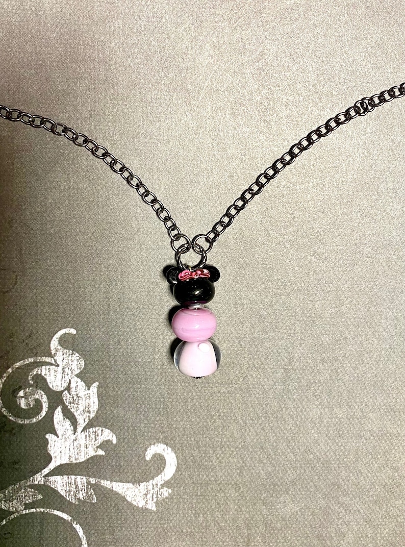 May include: A silver chain necklace with a black and pink bead charm. The charm is shaped like a cartoon character with a bow on its head.