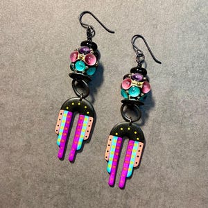May include: A pair of dangle earrings with black metal hooks and colorful glass beads. The bottom of the earrings feature a black and white geometric design with pink, blue, and yellow accents.