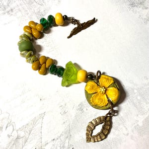 Puede incluir: Una pulsera artesanal con cuentas amarillas, verdes y color oliva. Incluye un dije de flor amarilla y un colgante en forma de hoja de bronce. El cierre es un dise&ntilde;o de hoja de bronce.