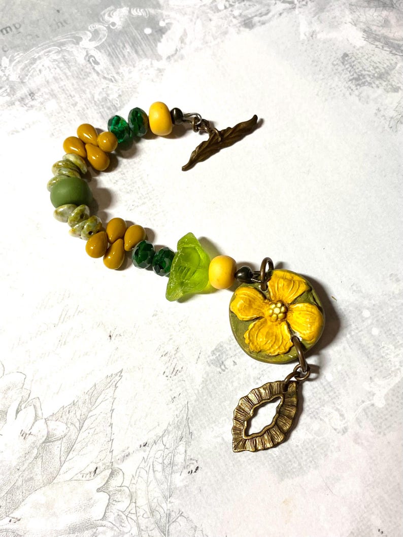 Puede incluir: Una pulsera artesanal con cuentas amarillas, verdes y color oliva. Incluye un dije de flor amarilla y un colgante en forma de hoja de bronce. El cierre es un dise&ntilde;o de hoja de bronce.