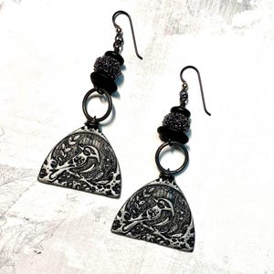 May include: A pair of silver earrings with a bird design. Each earring features a small, silver, triangular pendant with a bird perched on a branch. The pendant is attached to a silver ring and a series of black and silver beads.