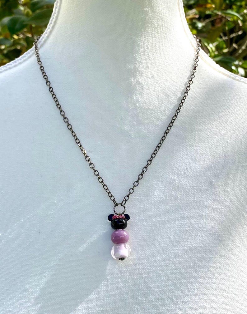 May include: A silver chain necklace with a pendant featuring three glass beads. The top bead is black with a pink bow, the middle bead is a light purple, and the bottom bead is white.