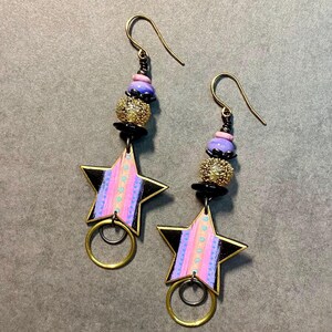 May include: A pair of dangle earrings with a star-shaped charm. The stars are painted black with pink and purple stripes. The earrings have gold hoops and are adorned with black, gold, and purple beads.