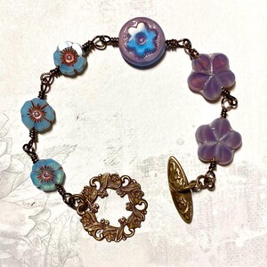 May include: A delicate bracelet featuring a mix of pastel blue and purple flower-shaped beads, connected by a copper chain. The bracelet has a decorative brass clasp with a floral design.