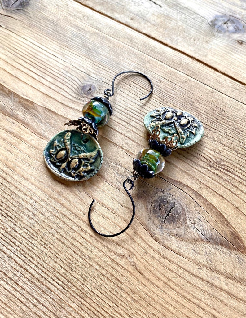 May include: A pair of green and gold ceramic earrings with dragonfly designs and black wire hooks. The earrings are strung with green and gold glass beads.