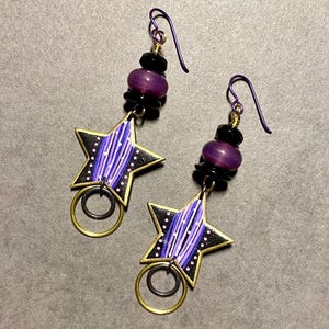 May include: Purple and black star-shaped earrings with gold rings and purple and black beads. The stars have a white and purple striped design.