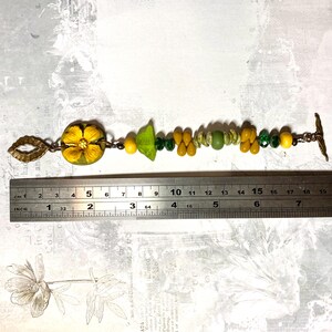 Puede incluir: Una pulsera hecha a mano con un dije de flor amarilla, cuentas verdes en forma de hoja y varias cuentas amarillas, verdes y transparentes. La pulsera mide aproximadamente 18 cm de largo, con un cierre y elementos decorativos de color bronce.