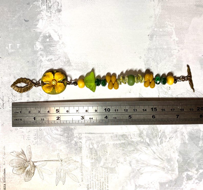 Puede incluir: Una pulsera hecha a mano con un dije de flor amarilla, cuentas verdes en forma de hoja y varias cuentas amarillas, verdes y transparentes. La pulsera mide aproximadamente 18 cm de largo, con un cierre y elementos decorativos de color bronce.
