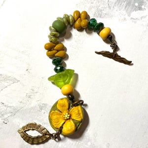 Puede incluir: Una pulsera artesanal con cuentas amarillas, verdes y oliva. Incluye un gran dije de flor amarilla y un cierre decorativo. Las cuentas est&aacute;n ensartadas con una cadena de metal. Una pieza de joyer&iacute;a &uacute;nica.