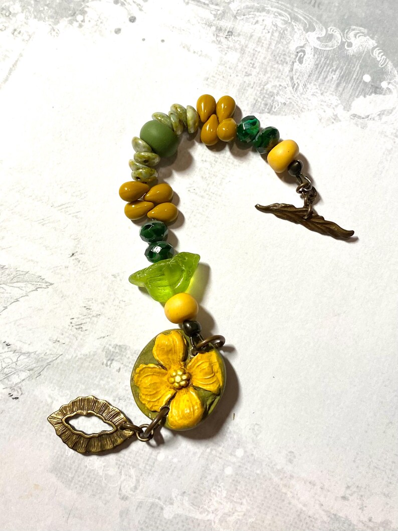 Puede incluir: Una pulsera artesanal con cuentas amarillas, verdes y oliva. Incluye un gran dije de flor amarilla y un cierre decorativo. Las cuentas est&aacute;n ensartadas con una cadena de metal. Una pieza de joyer&iacute;a &uacute;nica.
