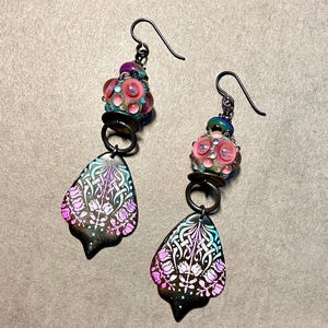 May include: A pair of dangle earrings with a black metal base and pink and blue glass beads. The earrings feature a teardrop-shaped pendant with a floral design.