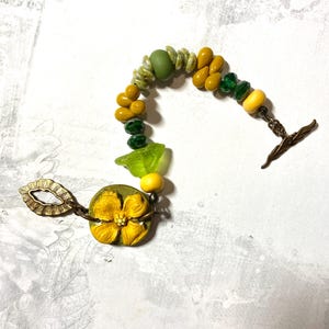 Puede incluir: Una pulsera hecha a mano con cuentas amarillas, verdes y transparentes. Cuenta con un gran dije de flor amarilla y un cierre de palanca de bronce. Las cuentas est&aacute;n ensartadas en una delicada cadena.