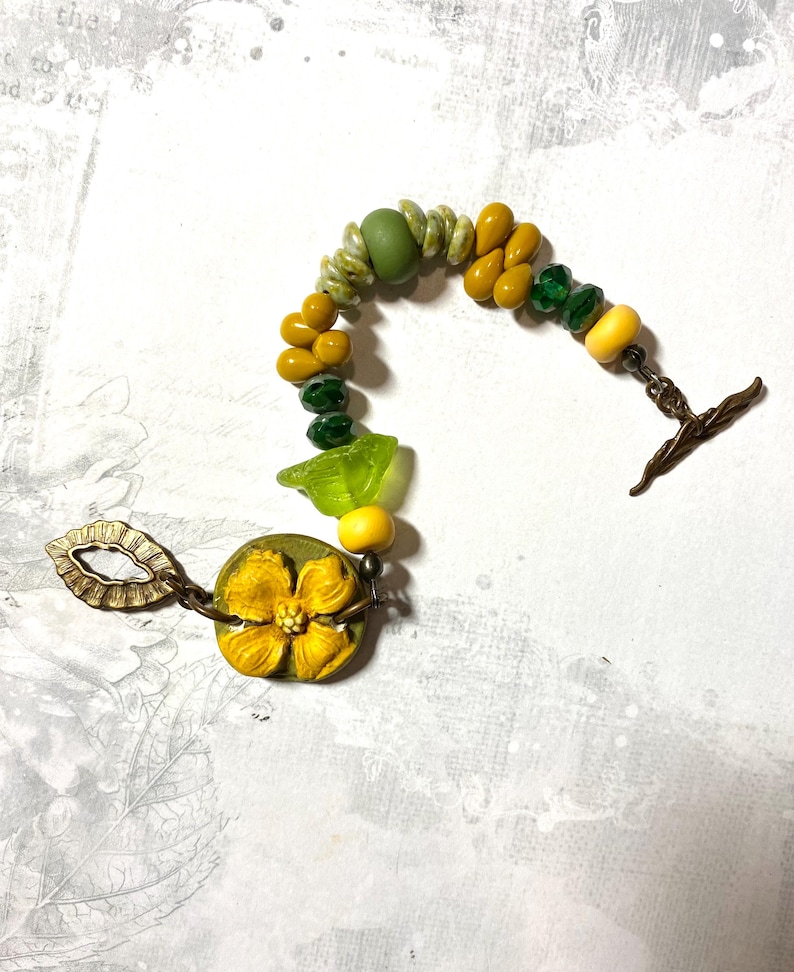 Puede incluir: Una pulsera hecha a mano con cuentas amarillas, verdes y transparentes. Cuenta con un gran dije de flor amarilla y un cierre de palanca de bronce. Las cuentas est&aacute;n ensartadas en una delicada cadena.