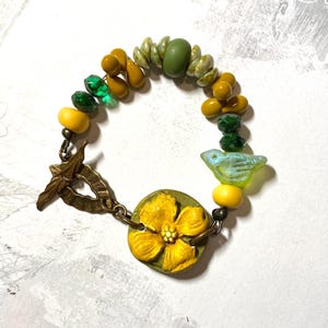 Puede incluir: Una pulsera artesanal con un dije de flor amarilla y un peque&ntilde;o dije de p&aacute;jaro azul. La pulsera incluye una variedad de cuentas en tonos amarillos, verdes y transparentes. El cierre es un cierre de palanca de color bronce.