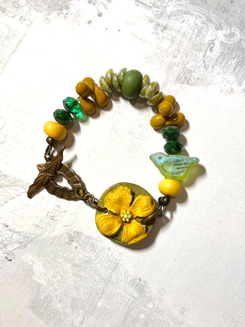 Puede incluir: Una pulsera artesanal con un dije de flor amarilla y un peque&ntilde;o dije de p&aacute;jaro azul. La pulsera incluye una variedad de cuentas en tonos amarillos, verdes y transparentes. El cierre es un cierre de palanca de color bronce.