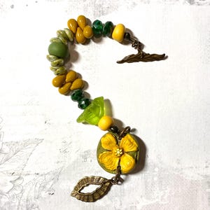 Puede incluir: Una pulsera artesanal con cuentas amarillas, verdes y oliva. Incluye un colgante de flor amarilla y un dije de hoja color bronce. El cierre es un cierre de palanca color bronce.