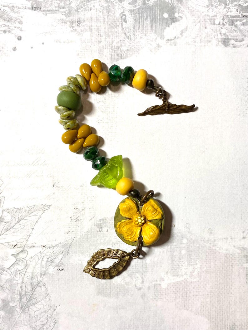 Puede incluir: Una pulsera artesanal con cuentas amarillas, verdes y oliva. Incluye un colgante de flor amarilla y un dije de hoja color bronce. El cierre es un cierre de palanca color bronce.