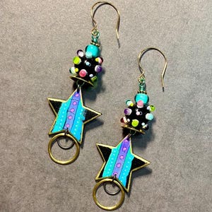 May include: A pair of dangle earrings with a black star charm, a turquoise and black bead cluster, and a gold ring. The star charm has a blue and purple striped pattern with white polka dots.