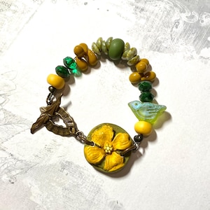 Puede incluir: Una pulsera artesanal con un dije de flor amarilla y una cuenta en forma de p&aacute;jaro. La pulsera incorpora cuentas amarillas, verdes y transparentes, conectadas por un cierre de tono bronce. La longitud de la pulsera es de aproximadamente 18 cm.