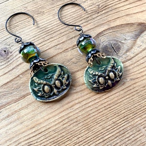 May include: A pair of green owl earrings with gold accents and iridescent green glass beads. The earrings are made of polymer clay and have a rustic, vintage look.