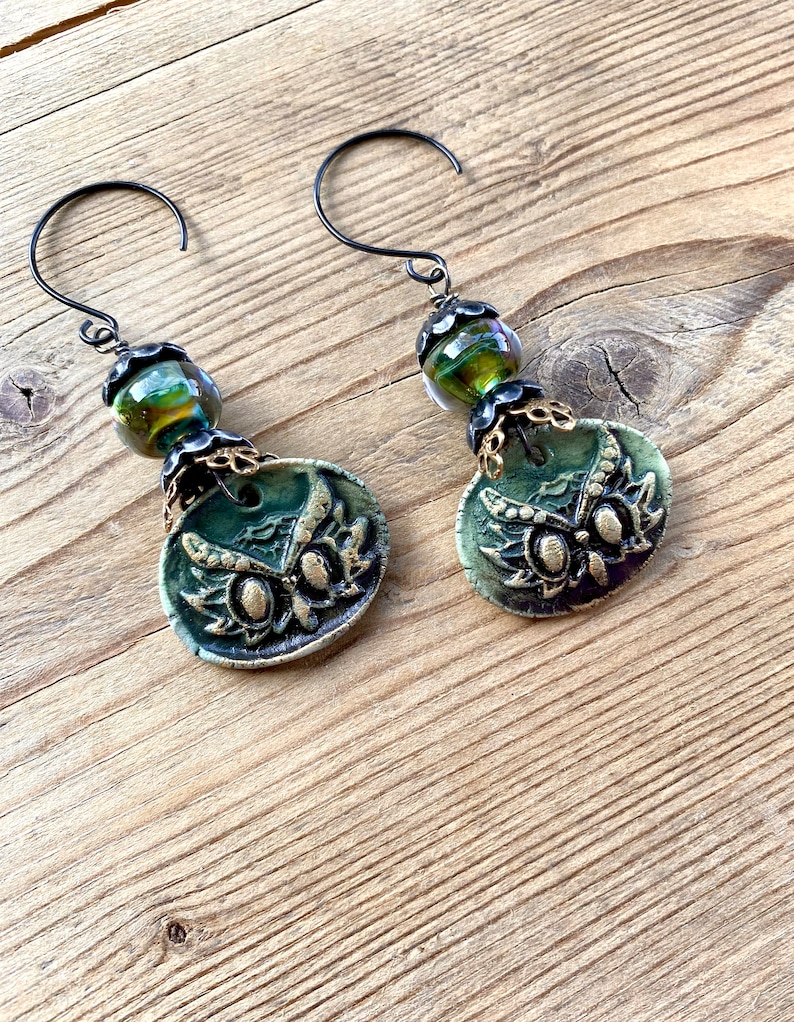 May include: A pair of green owl earrings with gold accents and iridescent green glass beads. The earrings are made of polymer clay and have a rustic, vintage look.